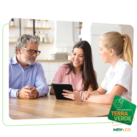 Um casal, composto por um homem mais velho de óculos e uma mulher mais jovem, conversa com uma consultora. A consultora, uma mulher loira, apresenta informações em um tablet, enquanto o casal olha para a tela com interesse e sorriso, em um ambiente claro e moderno. Abaixo o selo verde com o escrito " Programa Terra Verde - MRV&CO" 